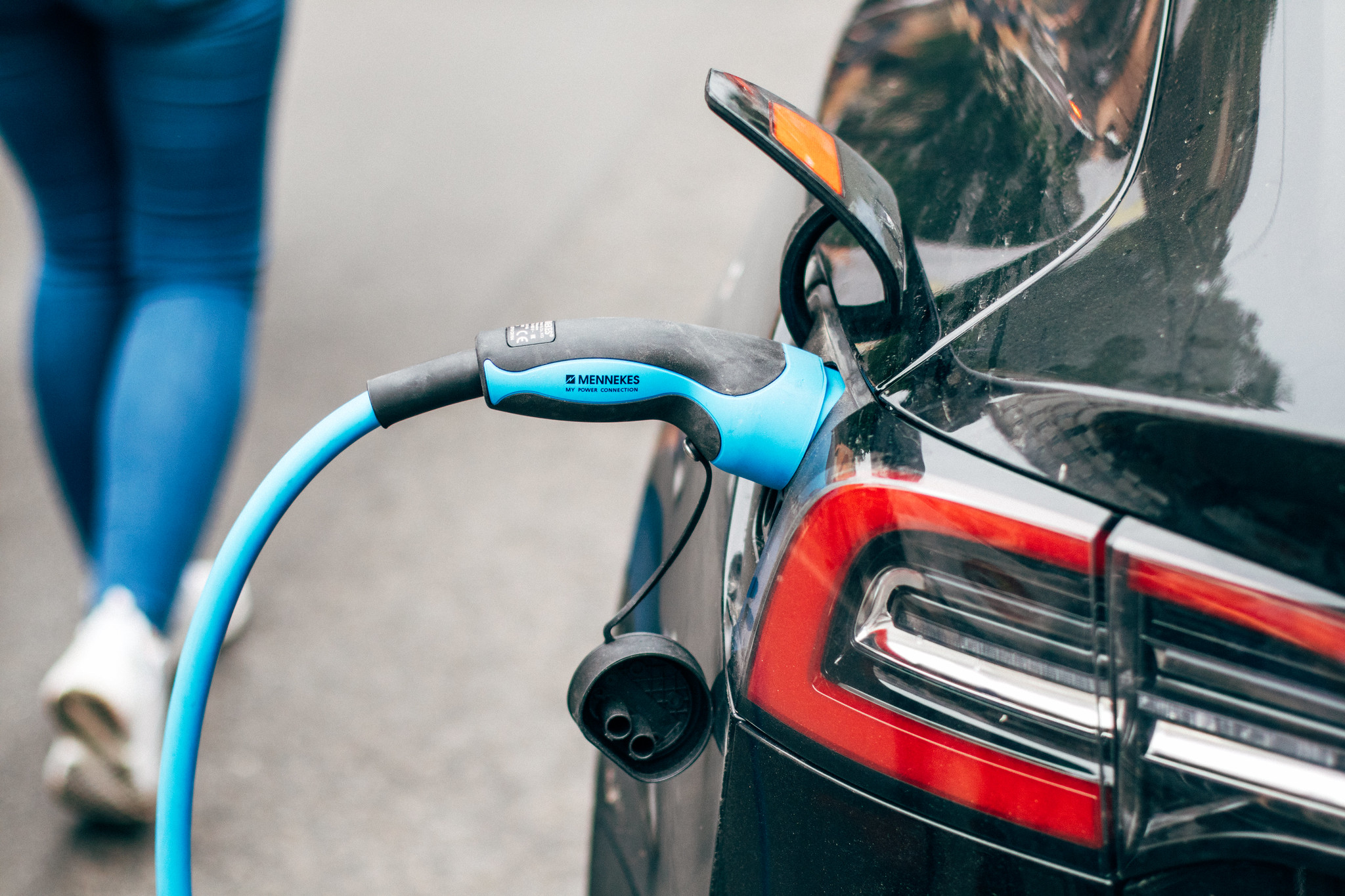 Gros plan d'une voiture électrique en train de se recharger, avec un câble de recharge bleu connecté à la prise de charge du véhicule. Une personne est partiellement visible en arrière-plan, marchant sur la route.
